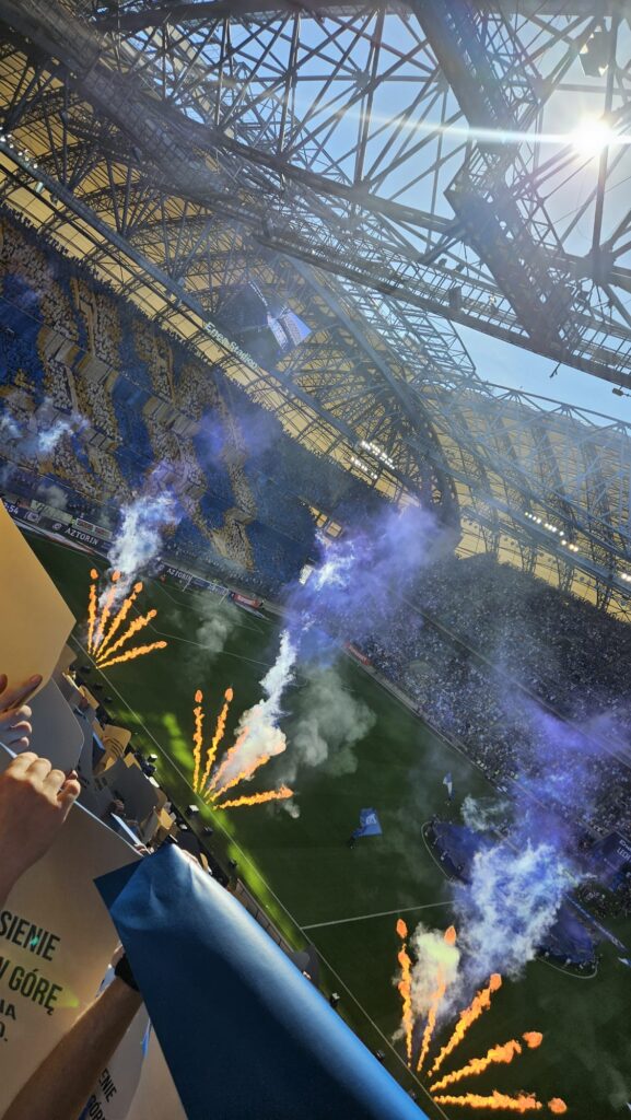 Wypełniony stadion z fanami trzymającymi znaki, żółte i niebieskie siedzenia, efekty dymu i ognia na boisku oraz światło słoneczne wpadające przez metalowy dach podczas tętniącego życiem meczu piłki nożnej lub wydarzenia.