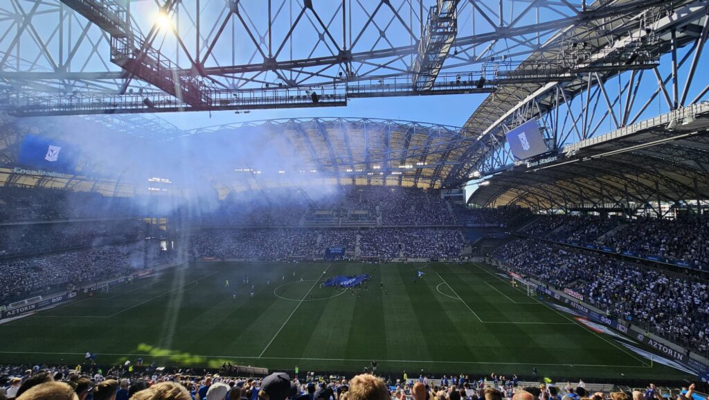 Na wypełnionym po brzegi stadionie z nowoczesnym dachem odbywa się mecz piłki nożnej. Kibice wypełniają trybuny, niektórzy wymachują flagami, a światło słoneczne wpada przez konstrukcję i mgiełka dymu unosi się nad boiskiem.