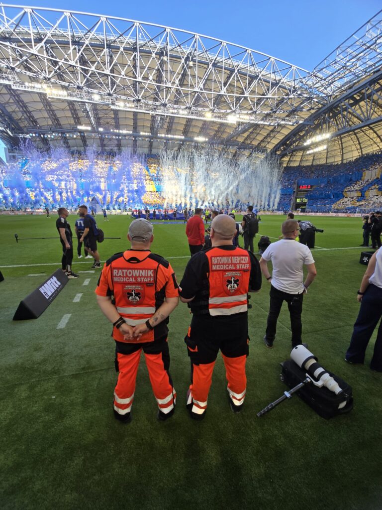 Dwóch pracowników medycznych w pomarańczowych strojach stoi na boisku piłkarskim, naprzeciwko stadionu wypełnionego kibicami, niebieskim i żółtym dymem oraz konfetti. Inni pracownicy i fotografowie są w pobliżu. Stadion ma zakrzywiony dach i jest jasno oświetlony.