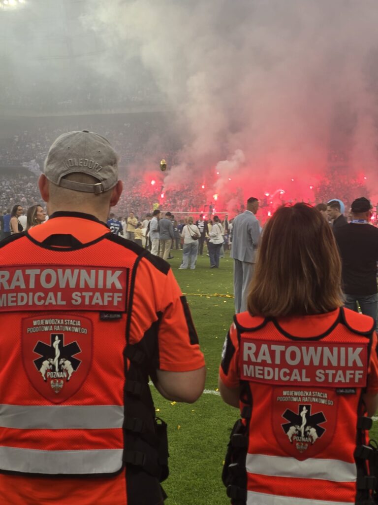 Dwóch pracowników medycznych w pomarańczowych kamizelkach stoi na boisku piłkarskim, naprzeciwko tłumu z czerwonymi racami i dymem w tle podczas wydarzenia na stadionie.
