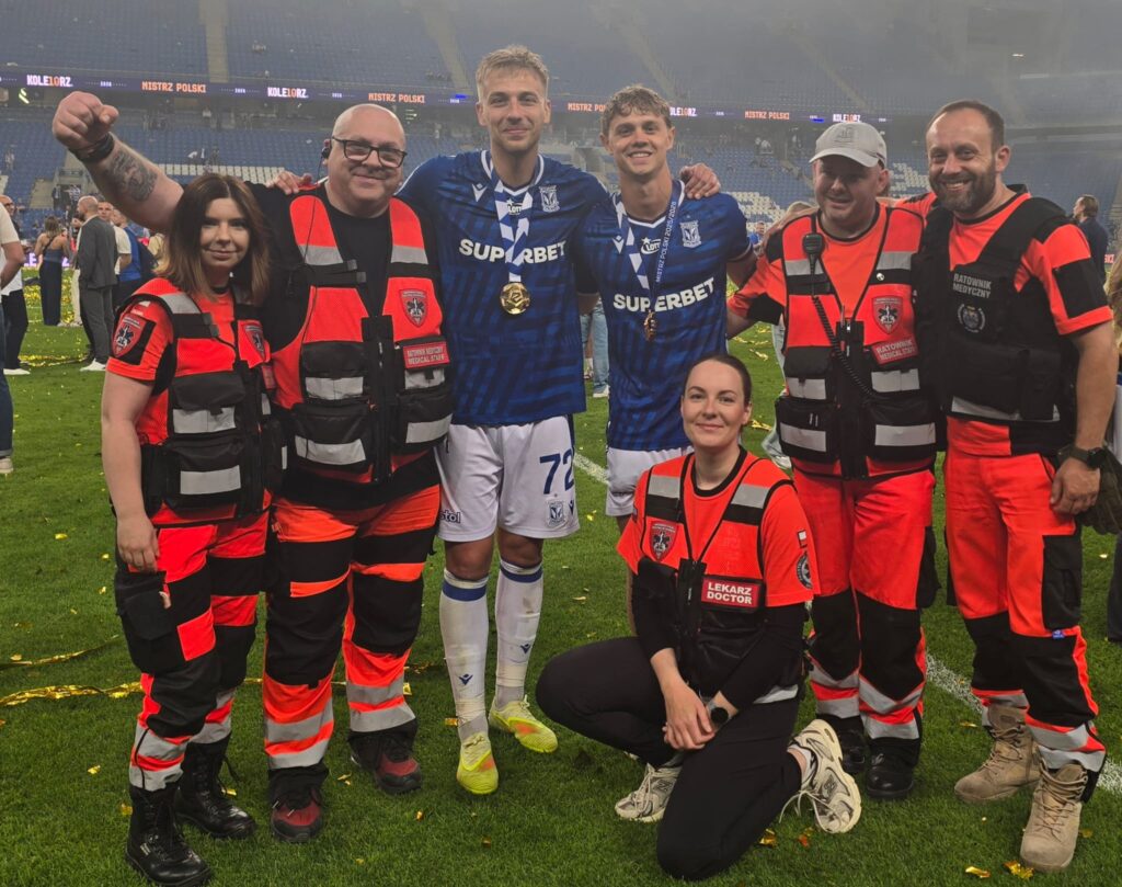 Grupa siedmiu osób, w tym dwóch piłkarzy w niebieskich koszulkach i pięciu medyków w czerwono-czarnych mundurach, pozuje razem na boisku piłkarskim po meczu, uśmiechając się do kamery.