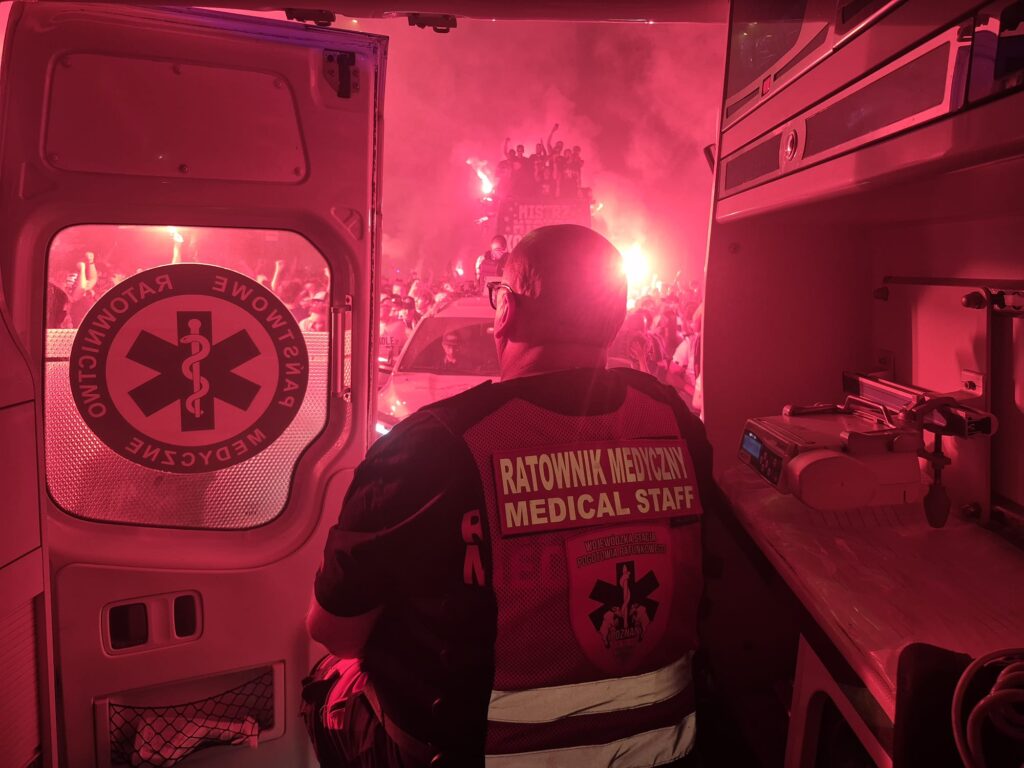 Członek personelu medycznego siedzi w otwartej karetce, obserwując tłum trzymający czerwone race w nocy. Scena na zewnątrz jest wypełniona dymem i czerwonym światłem, tworząc intensywną atmosferę.