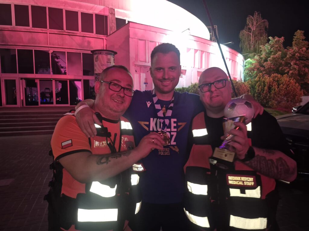 Trzech mężczyzn pozuje na zewnątrz w nocy; dwóch nosi pasujące pomarańczowo-czarne kamizelki personelu medycznego, jeden trzyma trofeum z piłką nożną na górze, a mężczyzna pośrodku uśmiecha się, mając na sobie niebieską koszulę i medal. Tło jest podświetlone na różowo.