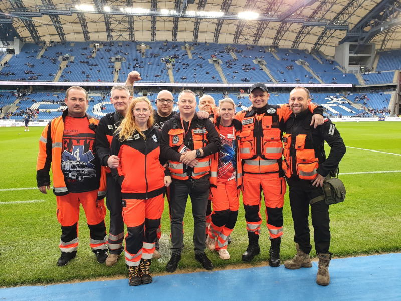 Grupa dziewięciu osób w pomarańczowych i czarnych strojach służb ratunkowych stoi uśmiechnięta na boisku piłkarskim z miejscami siedzącymi na stadionie i widzami w tle.