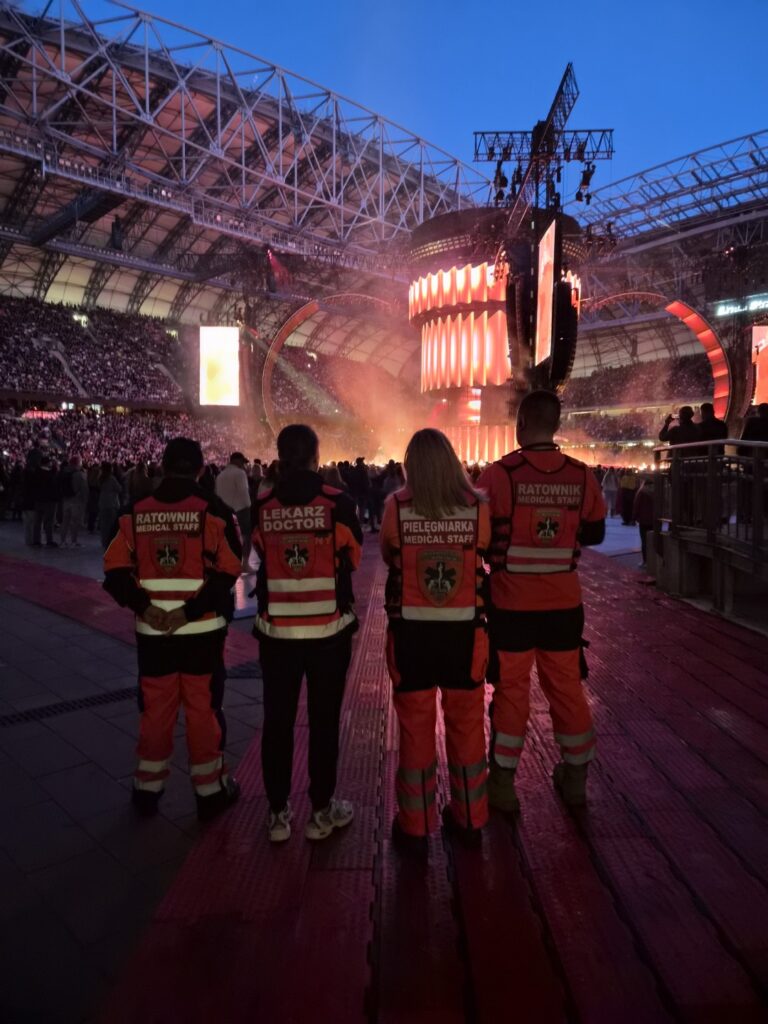 Czterech ratowników medycznych w czerwonych uniformach stoi na skraju stadionu, patrząc w stronę jasno oświetlonej sceny koncertowej i ogromnego tłumu zgromadzonego pod otwartym dachem stadionu o zmierzchu.