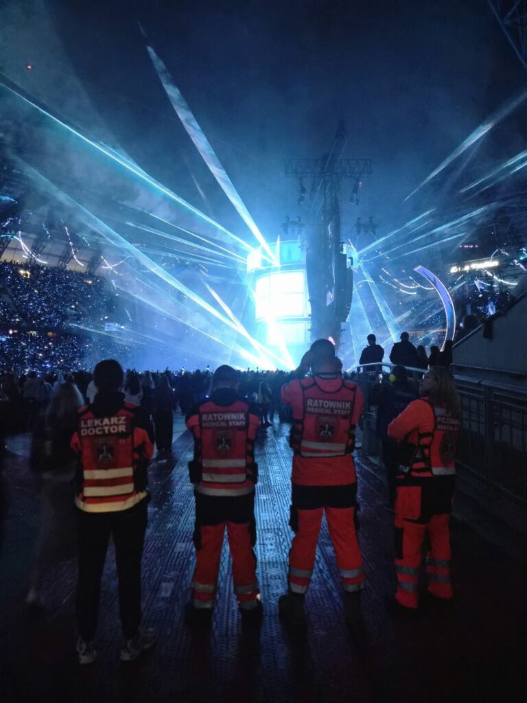 Czterech ratowników w odblaskowych mundurach stoi twarzą do jasno oświetlonej sceny koncertowej z niebieskimi światłami laserowymi, czuwając nad ogromnym tłumem zgromadzonym na stadionie.