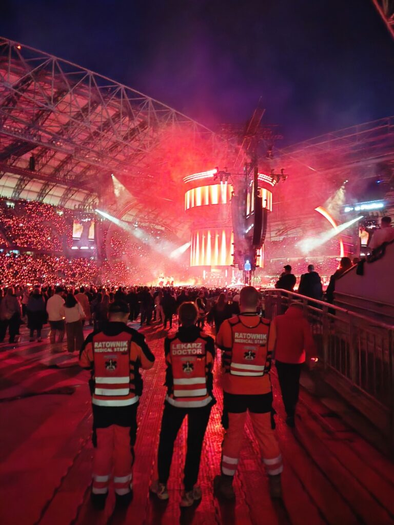 Trzech ratowników w czerwonych mundurach stoi twarzą do zatłoczonego stadionu, na którym podczas nocnego koncertu zgromadził się tłum ludzi, a scenę oświetlają jaskrawoczerwone reflektory.