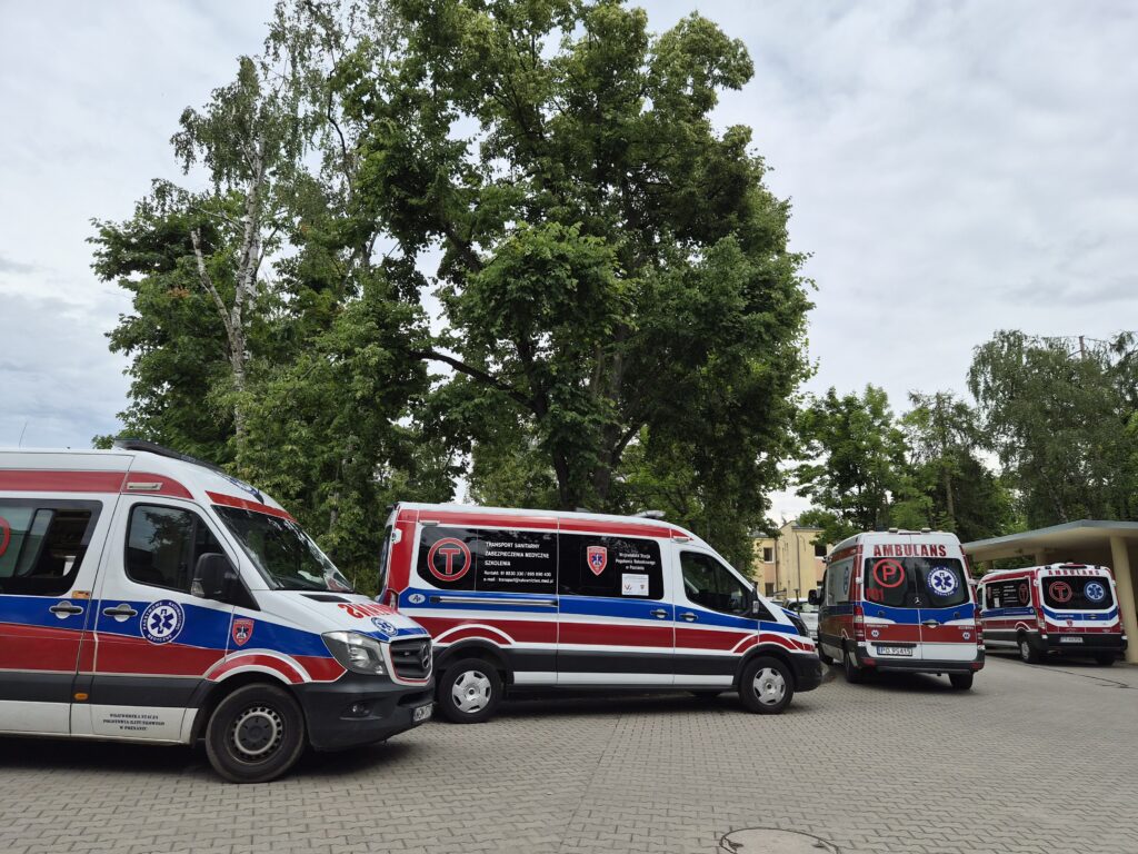 Cztery karetki pogotowia z czerwono-biało-niebieskimi oznaczeniami stoją na utwardzonym placu w pobliżu wysokich zielonych drzew pod zachmurzonym niebem. Pojazdy są ustawione obok siebie, skierowane w różnych kierunkach.