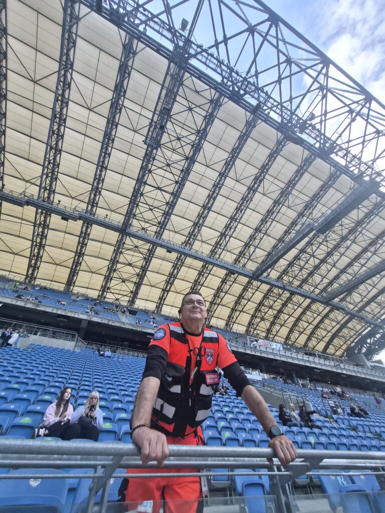 Mężczyzna w pomarańczowej kamizelce odblaskowej stoi przy barierce na prawie pustym stadionie z dużym, łukowatym dachem. W tle, pod częściowo zachmurzonym niebem, na niebieskich siedzeniach siedzi kilka osób.