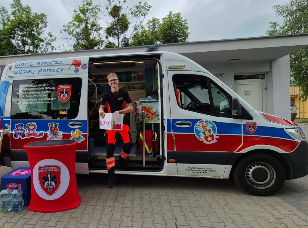 Uśmiechnięty ratownik medyczny w pomarańczowo-czarnym mundurze stoi przy otwartych drzwiach udekorowanej karetki, trzymając w ręku torbę z prezentami. Obok pojazdu stoi czerwony stolik z butelkami wody i ulotkami.