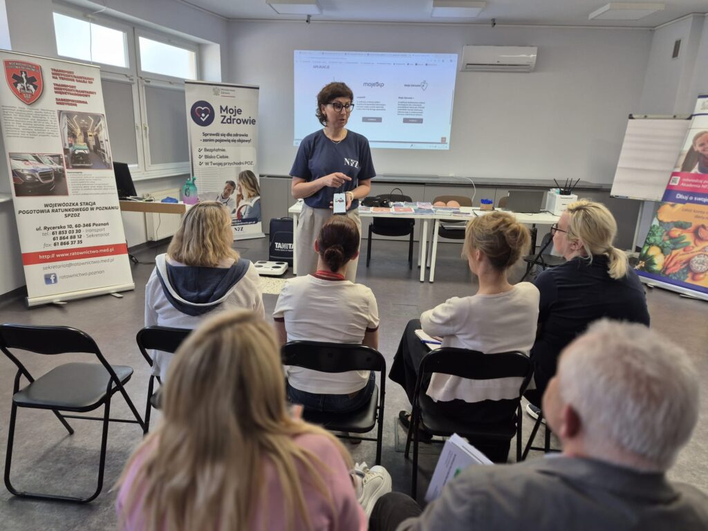 Kobieta stoi z przodu klasy, prowadząc prezentację dla siedzącej grupy dorosłych. Za nią znajdują się banery, ekran projekcyjny z diagramami i materiały informacyjne na stołach.