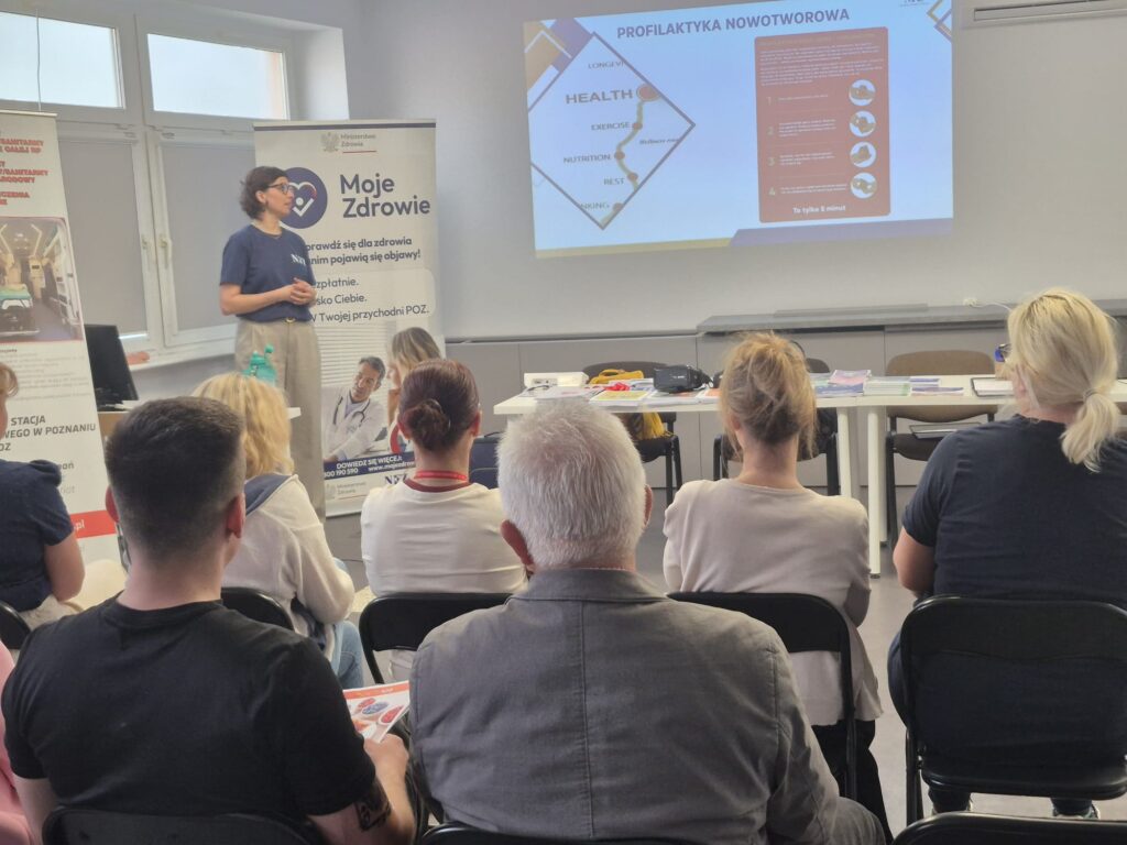 Kobieta prowadzi prezentację na temat profilaktyki raka dla publiczności w klasie. Widoczny jest slajd PowerPoint i banery zdrowotne w języku polskim. Uczestnicy siedzą i słuchają, niektórzy czytają broszury.