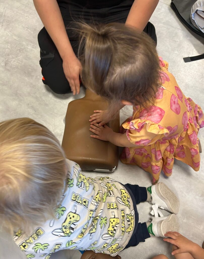 Dwoje małych dzieci wykonuje uciski klatki piersiowej na manekinie do nauki resuscytacji, pod okiem osoby dorosłej. Dziewczynka ma na sobie r&oacute;żową sukienkę, a chłopiec ma blond włosy i koszulkę w wzory.