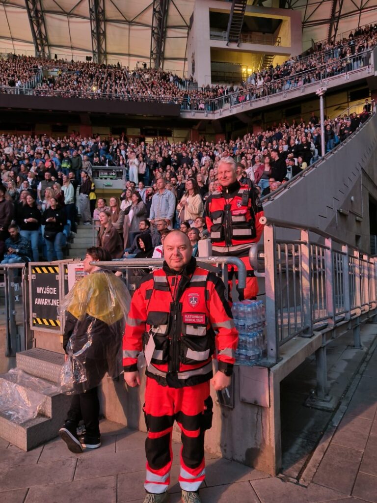 Dwóch ratowników w czerwono-czarnych mundurach stoi przy barierce na zatłoczonym stadionie, a w tle widać wielu widzów siedzących i stojących podczas imprezy.
