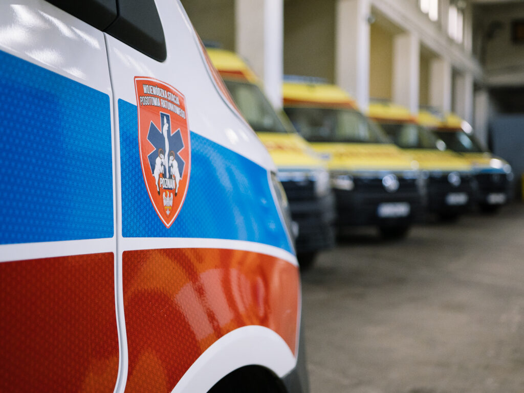 Zbliżenie karetki pogotowia z czerwonym, niebieskim i białym wzorem oraz plakietką pogotowia ratunkowego, zaparkowanej w garażu z kilkoma żółtymi karetkami widocznymi w tle.