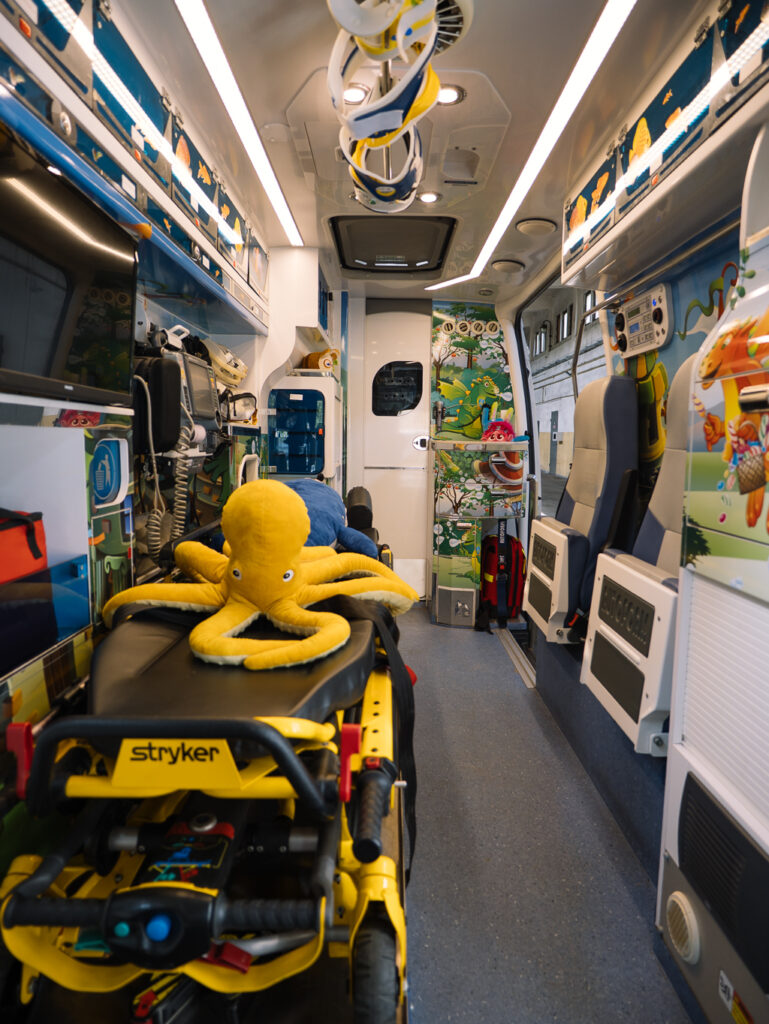 Wnętrze jasno udekorowanego ambulansu zawiera kolorowe ilustracje zwierząt i ż&oacute;łte nosze z pluszową ż&oacute;łtą ośmiornicą na g&oacute;rze, tworząc przyjazne dla dzieci środowisko.