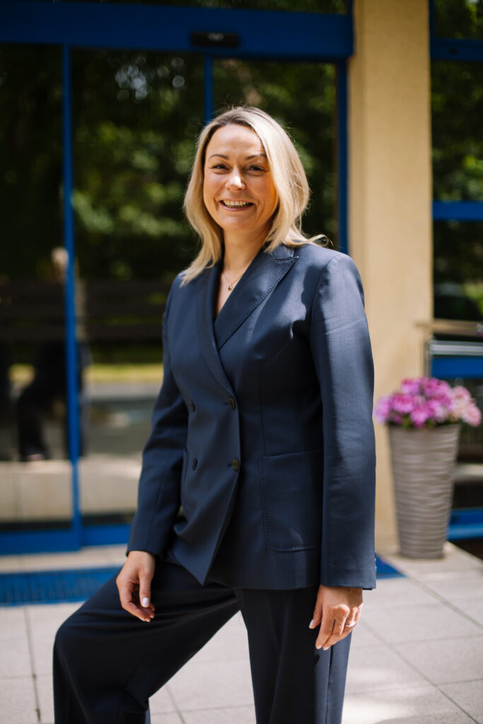 Kobieta o blond włosach, ubrana w granatowy garnitur, stoi na świeżym powietrzu przed budynkiem z dużymi oknami i doniczkową rośliną o fioletowych kwiatach, uśmiechając się.