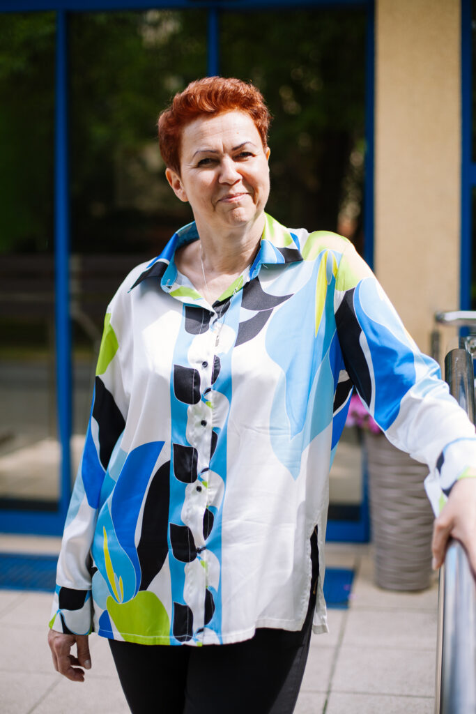 Kobieta o krótkich rudych włosach stoi na zewnątrz, ubrana w kolorową koszulę w wzory z guzikami, z motywami w kolorach niebieskim, zielonym, żółtym i czarnym; uśmiecha się lekko, opierając jedną rękę na poręczy.