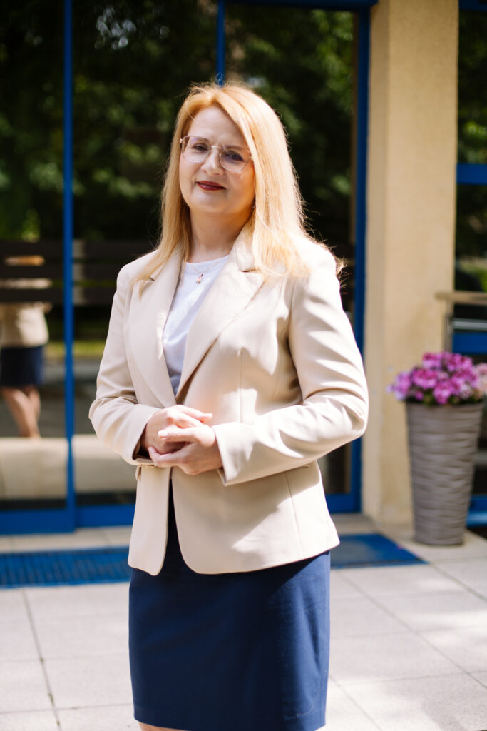 Kobieta o długich blond włosach, w okularach, beżowej marynarce i granatowej spódnicy, stoi na zewnątrz przy wejściu do budynku z szklanymi drzwiami i dużą rośliną doniczkową o fioletowych kwiatach.
