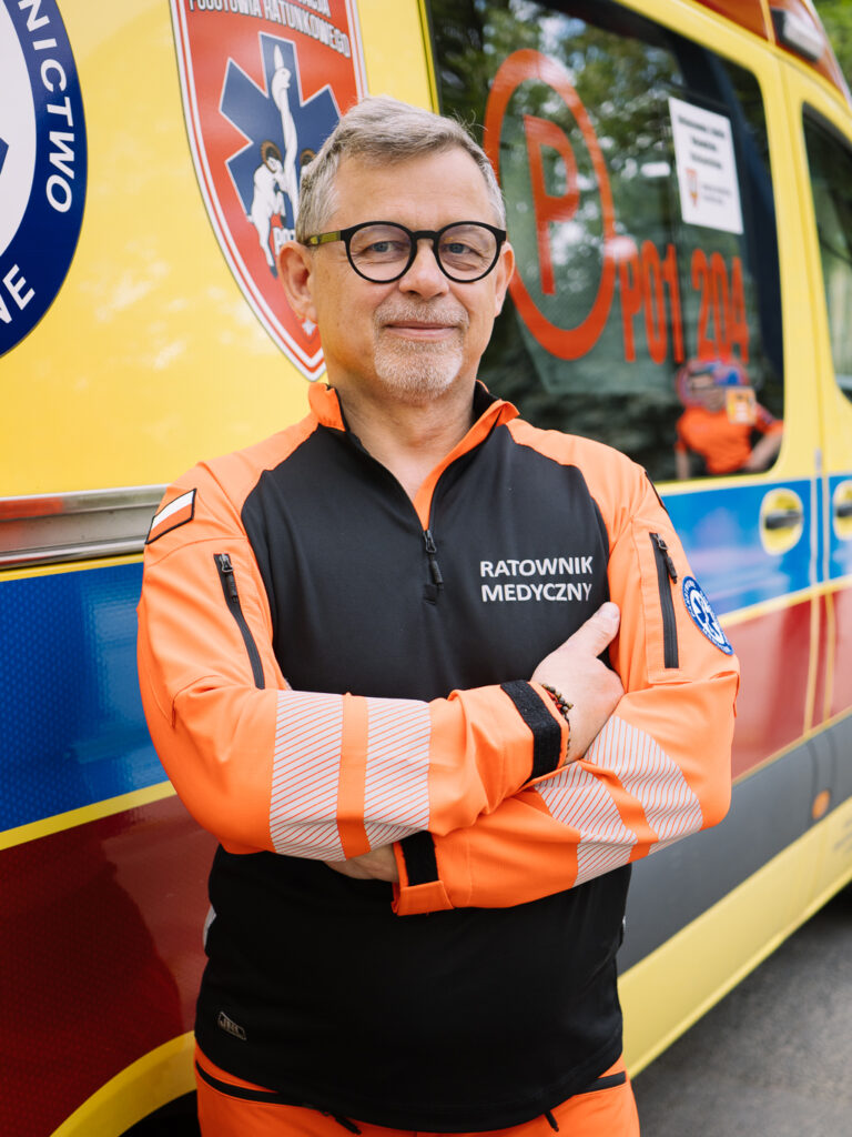 Ratownik medyczny w średnim wieku, ubrany w pomarańczowo-czarny mundur, stoi ze skrzyżowanymi rękami przed karetką. Na mundurze widnieje napis „Ratownik Medyczny”, co w języku polskim oznacza ratownika medycznego.