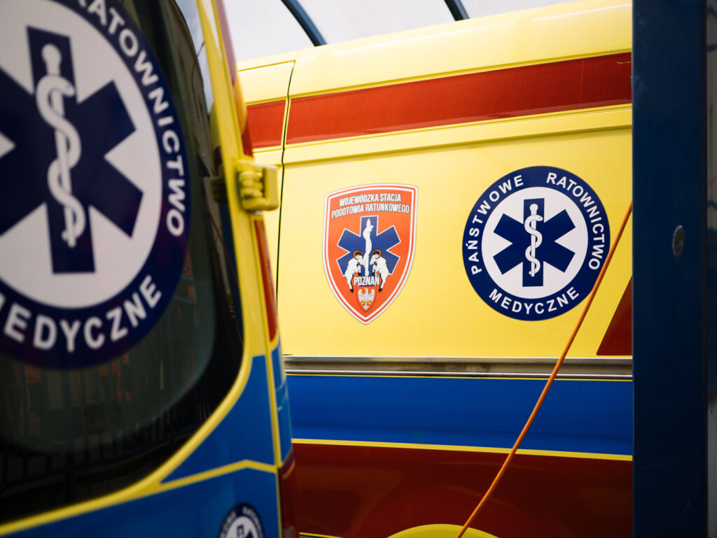 Zbliżenie na polską karetkę pogotowia z widocznymi symbolami i logo służb ratownictwa medycznego, w tym napisem „Państwowe Ratownictwo Medyczne” oraz czerwoną tarczą z białym orłem i niebieską gwiazdą życia.