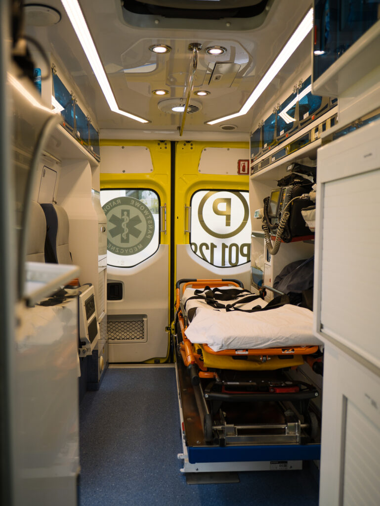 Wnętrze ambulansu ze sprzętem medycznym, pustymi noszami, biało-żółtymi ścianami i symbolami awaryjnymi widocznymi na tylnych drzwiach.