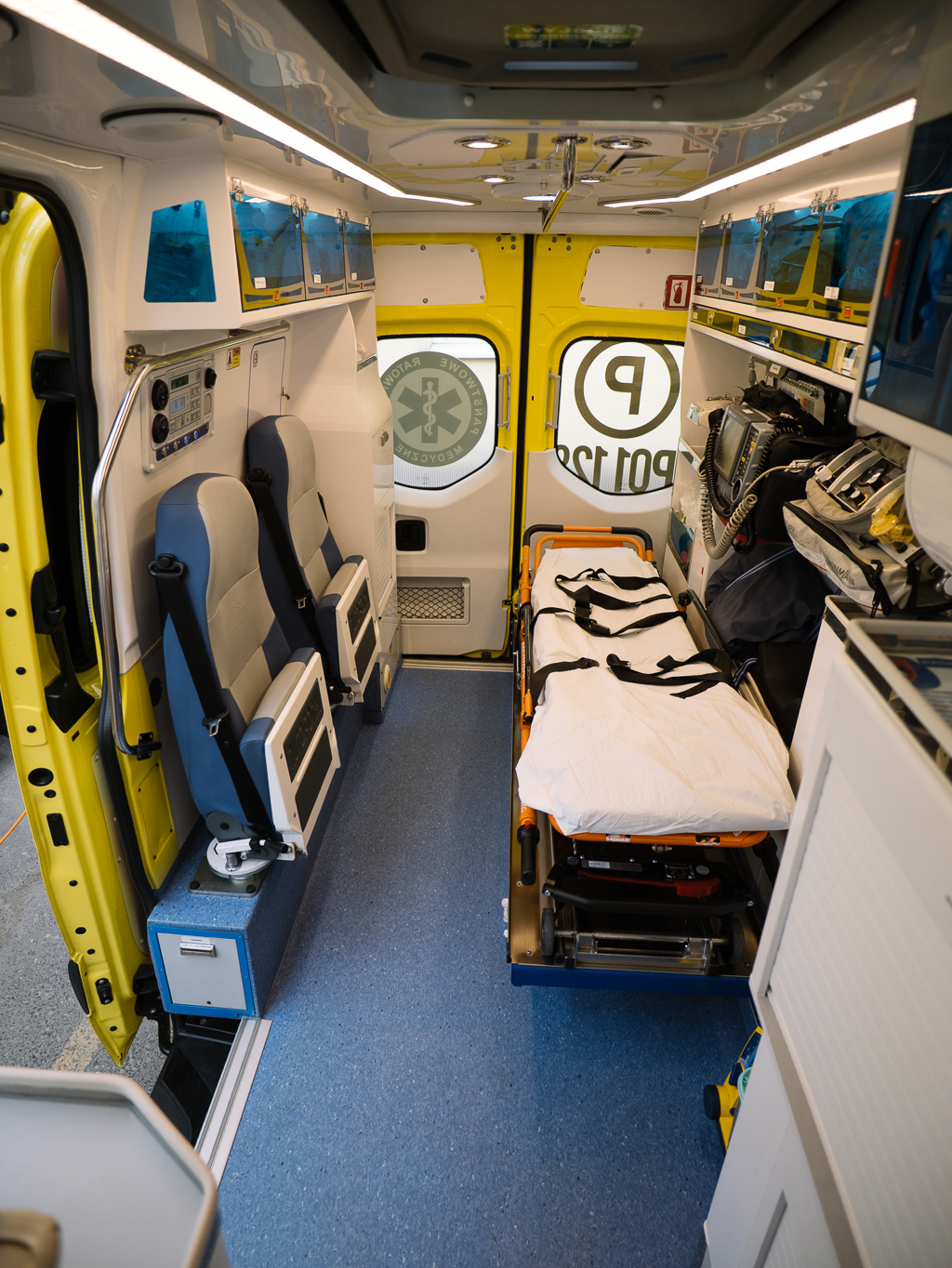 Wnętrze ambulansu z dwoma fotelami, sprzętem medycznym, szafkami i noszami z białym prześcieradłem, gotowe do transportu pacjenta. Tylne drzwi są częściowo otwarte, odsłaniając oznaczenia awaryjne.