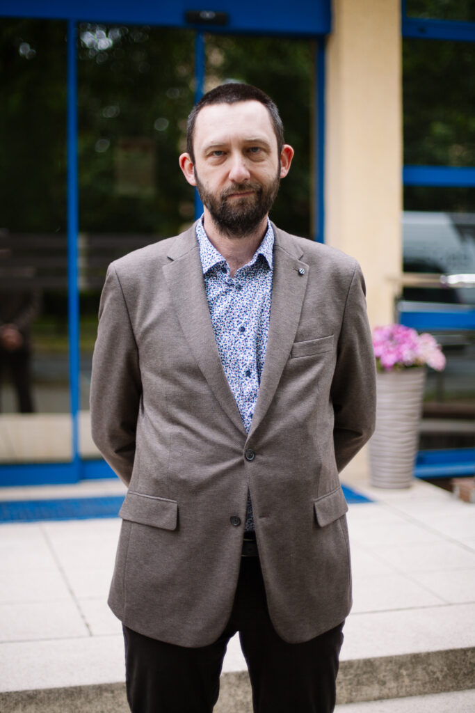 Mężczyzna z brodą, ubrany w szarą marynarkę i wzorzystą koszulę, stoi na zewnątrz przed budynkiem z niebieskimi ramami okiennymi, obok którego stoi roślina w doniczce, i patrzy prosto w obiektyw.