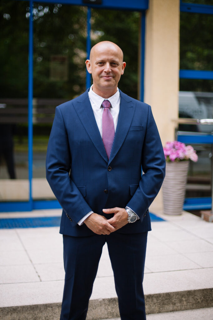 Łysy mężczyzna w niebieskim garniturze i różowym krawacie stoi przed budynkiem z dużymi oknami, a przy wejściu stoi doniczkowa roślina. Uśmiecha się i trzyma ręce złożone przed sobą.