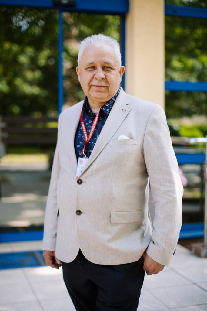 Starszy mężczyzna o krótkich białych włosach, ubrany w jasnobeżową marynarkę, granatową koszulę oraz czerwoną smycz z identyfikatorem, stoi na zewnątrz przed budynkiem z dużymi oknami, a w tle widać zieleń.
