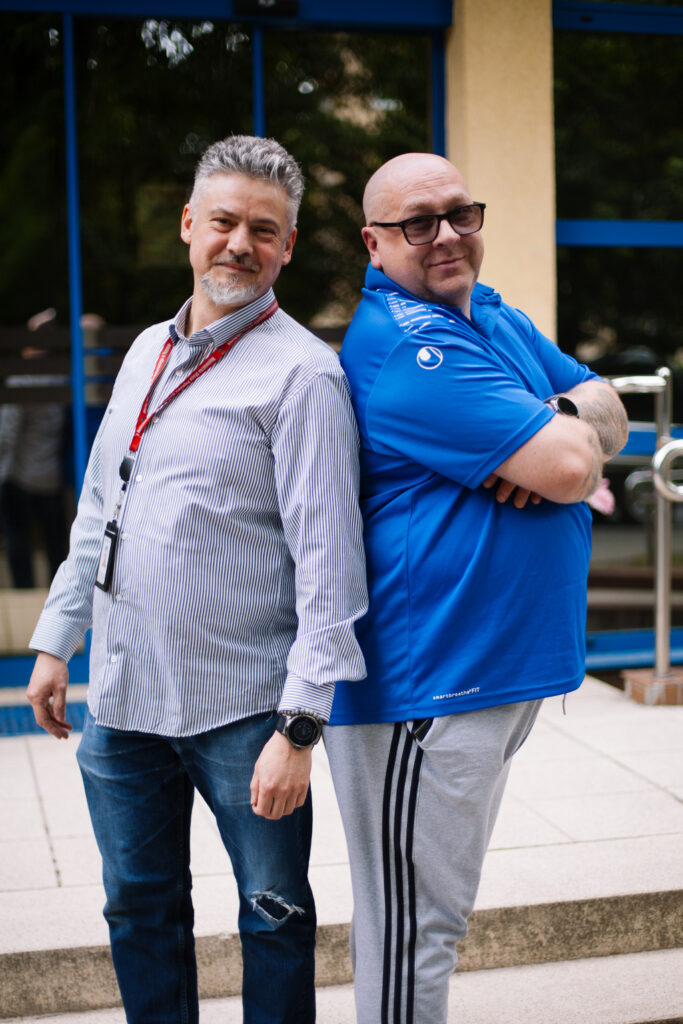 Dwóch mężczyzn stoi plecami do siebie na zewnątrz, uśmiechając się. Jeden ma na sobie koszulę w paski, dżinsy i smycz; drugi nosi okulary przeciwsłoneczne, niebieską koszulkę polo i szare spodnie dresowe. Wyglądają na zrelaksowanych i przyjaznych.