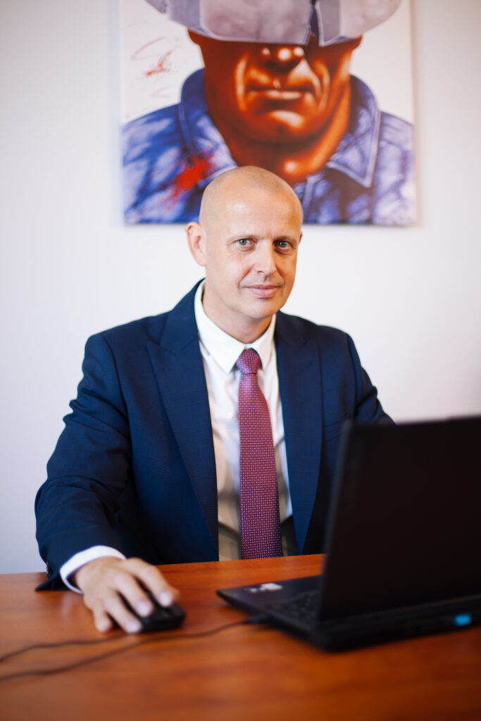 Łysy mężczyzna w niebieskim garniturze i różowym krawacie siedzi przy biurku i korzysta z laptopa, a na ścianie za nim znajduje się obraz przedstawiający osobę w kasku.
