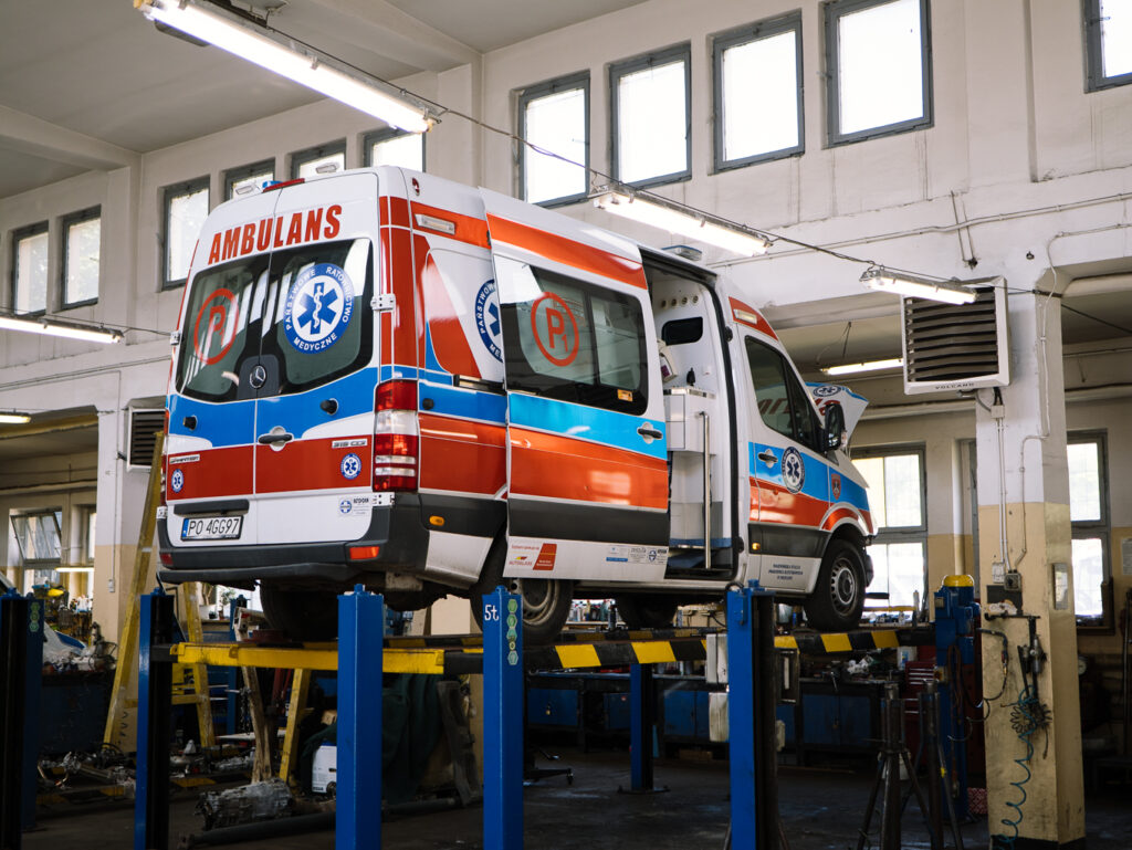 Czerwono-biały ambulans jest podniesiony na podnośniku hydraulicznym wewnątrz dobrze oświetlonego warsztatu samochodowego, z narzędziami i sprzętem widocznymi w tle.