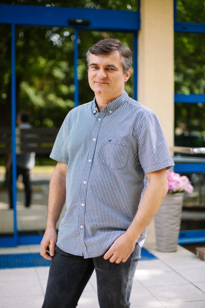 Mężczyzna w koszuli w kratkę z krótkim rękawem i ciemnych dżinsach stoi na zewnątrz przed szklanymi drzwiami; w tle widać zieleń i dużą roślinę doniczkową.