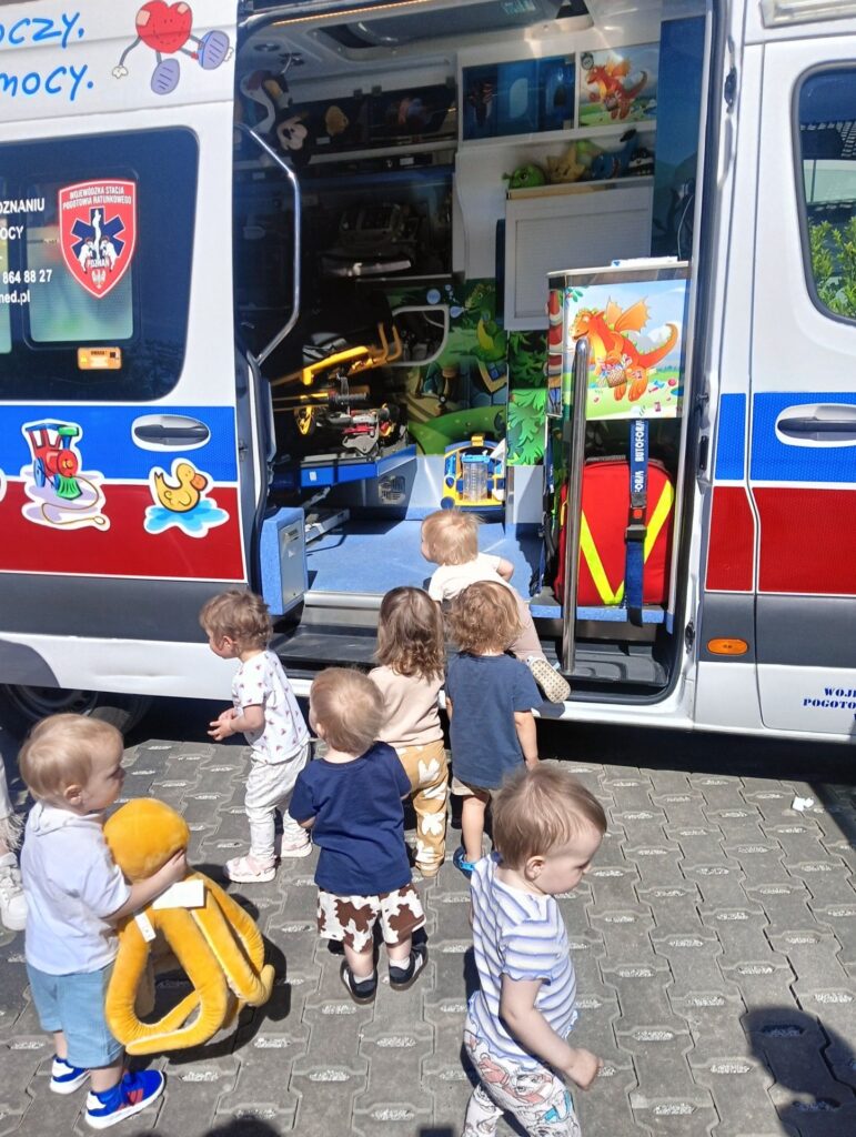 Grupa małych dzieci stoi przed otwartą karetką, zaglądając do środka, gdzie widać zabawki i kolorowe dekoracje. Jedno z dzieci trzyma w rękach dużą pluszową zabawkę. Scena rozgrywa się na świeżym powietrzu w słoneczny dzień.